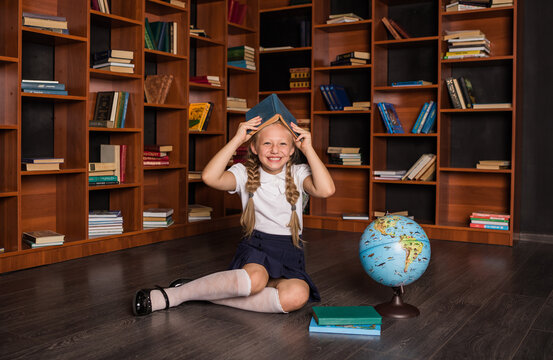 Happy girl pupil in school uniform sitting with a book on her head and laughing in class
