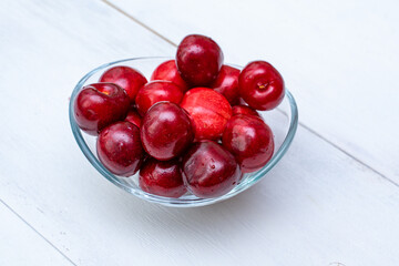 Glass vase in the form of a bowl with ripe cherries stands on a white wooden background.