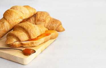 Fresh croissants lie on a wooden Board, next to a spoon with apricot jam. Light grey background, space for text
