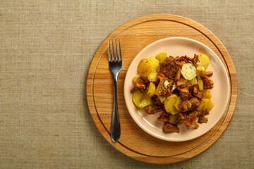 A plate of fried potatoes with larva mushrooms on a linen tablecloth on a round set and a fork.