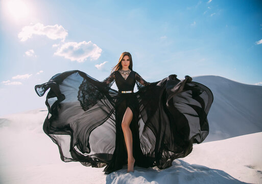 Beautiful sexy woman queen in black clothes. Girl fashion model. long silk dress fabric flies in wind, luxury glamorous elegant goddess shows bare legs in cut of skirt. background white sand blue sky