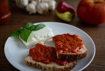Multigrain bread slice spread with traditional bulgarian tomato meal lutenica on focus with tomatoes, feta cheese, basil, peppers and garlic on background.
