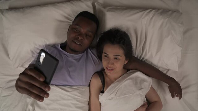 Top view of multi-ethnic couple lying in bed at night and talking with friends or relatives by video call. White woman waving hand, black man showing peace-sign