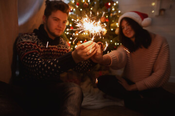 Obraz na płótnie Canvas Happy New Year. Happy couple holding fireworks under christmas tree with lights. Young family with burning sparklers celebrating together in festive dark room.
