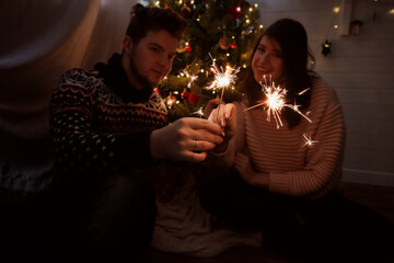 Obraz na płótnie Canvas Happy couple holding fireworks under christmas tree with lights. Young family with burning sparklers celebrating together in festive dark room. Happy New Year