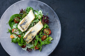 Traditional fried skrei cod fish filet with Italian lettuce and tomatoes in a lemon coconut sauce offered as top view in a modern design plate with copy space right