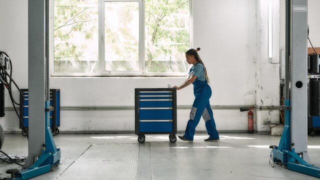 African American Woman, Professional Female Mechanic Pulling, Carrying Tool Box Cart In Auto Repair Shop. Car Service, Maintenance And People Concept