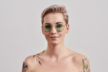 Portrait of a young attractive half naked tattooed woman wearing sunglasses and looking at camera isolated over light background