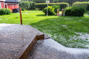 Obraz premium View at wooden terrace, stone path and green garden with hail stones during hailstorm from sky with sunlight