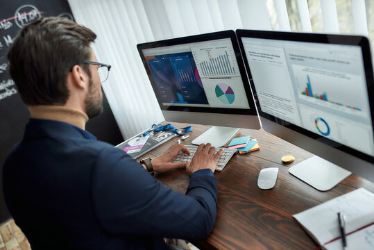 Preparing report. Rear view of focused businessman or financial analyst working in the modern office, looking at graphs and charts on pc screen, analyzing statistical data