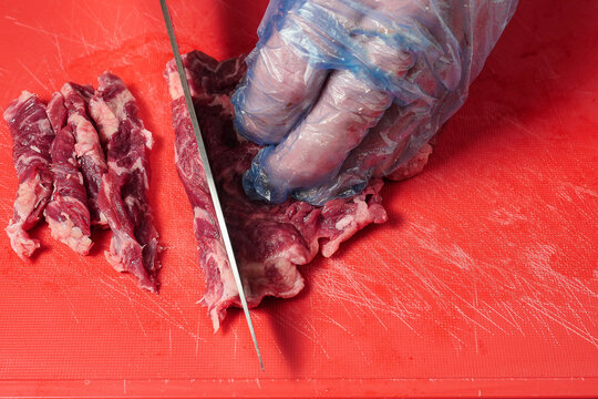 Butcher cutting fresh juicy rib eye steak into fine strips for stir fry on a red plastic cutting board with a sharp knife and holding meat with hand in blue glove. Meat industry concept.