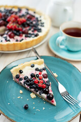 Piece of blueberry open pie on blue plate with cup of black tea and white tea pot on bright painted wood background