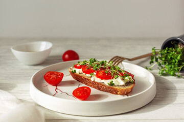 Tasty sandwich with cherry tomatoes, micro greens and soft cheese on wholegrain bread on light wooden background. Healthy breakfast or lunch concept.