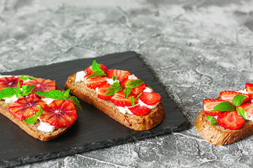 Tasty sandwiches with strawberry, blood orange, soft cheese and basil leaves on roasted wholegrain bread served on black feed board. Healthy delicious breakfast or lunch concept.
