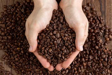 A handful or scattering of roasted coffee beans in handsome male hands over a wooden table. Coffee shop, wallpaper. Place for text