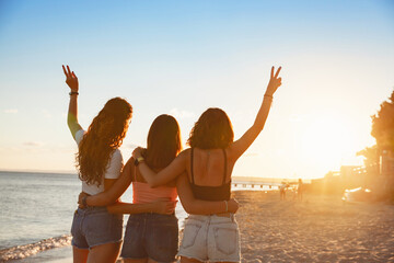 Group of three beautiful girls staying and hugging with raised arms on the beach. Travel or sea vacations concept. Three young happy women in the summer. The view from the back.