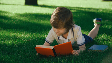 Closeup view of young kid lying and relaxing on green grass. Boy reading paper book, turning pages outside in summer or spring, meadow. 