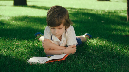 Closeup view of young kid lying and relaxing on green grass. Boy reading paper book, turning pages outside in summer or spring, meadow. 
