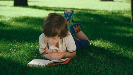Closeup view of young kid lying and relaxing on green grass. Boy reading paper book, turning pages outside in summer or spring, meadow. 