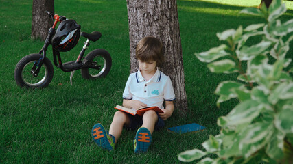 A boy is sitting under a big tree on the green grass and reading a book.