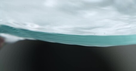 Macro shot of the edge of a glass filled with water. Water moves in rhythmic patterns. Glass gets emptied at the end of the shot. - Powered by Adobe