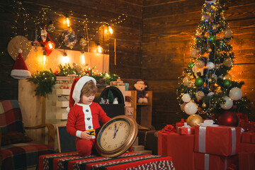 Smiling little Santa helper boy looks at clock and waiting for New Year midnight at luxury...