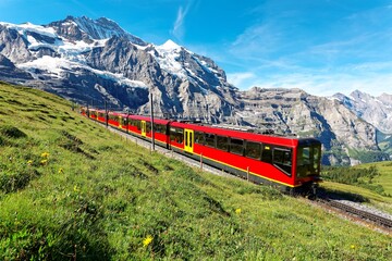 Fototapeta na wymiar A cogwheel train travels on the railway from Jungfraujoch to Kleine Scheidegg on the green grassy hillside with Jungfrau & Monch mountains in background, in Bernese Highlands, Switzerland
