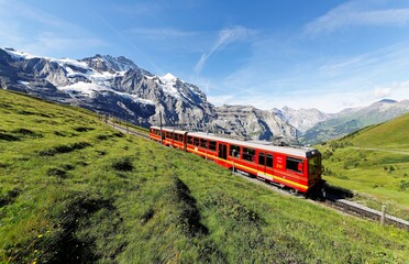 Fototapeta na wymiar Tourists traveling on a cogwheel train of the famous Jungfrau Railway from Jungfraujoch (Top of Europe) to Kleine Scheidegg on a green grassy hillside under sunny sky, in Bernese Oberland, Switzerland