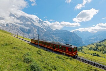 Naklejka na ściany i meble A cogwheel train travels on the railway from Jungfraujoch to Kleine Scheidegg on the green grassy hillside with Jungfrau & Monch mountains in background, in Bernese Highlands, Switzerland