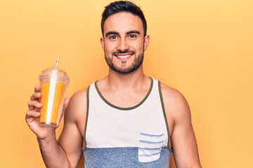 Handsome man with beard wearing sleeveless t-shirt drinking glass of healthy orange juice looking positive and happy standing and smiling with a confident smile showing teeth