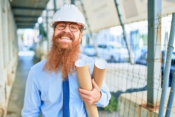 Young redhead architect man with long beard wearing hardhat smiling happy. Standing with smile on face holding blueprints at street of city.