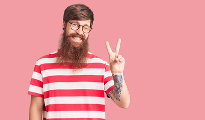 Handsome young red head man with long beard wearing casual clothes smiling with happy face winking at the camera doing victory sign. number two.