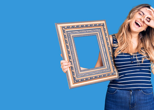 Young caucasian woman holding empty frame smiling happy doing ok sign with hand on eye looking through fingers
