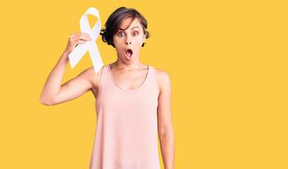 Beautiful young woman with short hair holding cancer awareness white ribbon scared and amazed with open mouth for surprise, disbelief face