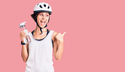Beautiful young woman with short hair wearing bike helmet and holding water bottle pointing thumb up to the side smiling happy with open mouth