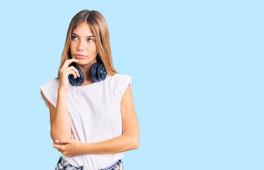 Beautiful caucasian woman with blonde hair listening to music wearing headphones serious face thinking about question with hand on chin, thoughtful about confusing idea