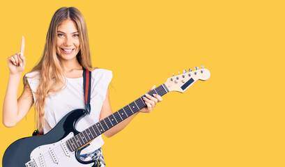Fototapeta premium Beautiful caucasian woman with blonde hair playing electric guitar surprised with an idea or question pointing finger with happy face, number one