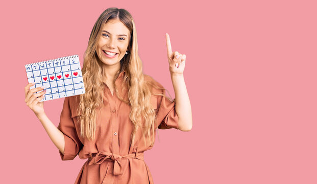 Beautiful caucasian woman with blonde hair holding heart calendar surprised with an idea or question pointing finger with happy face, number one