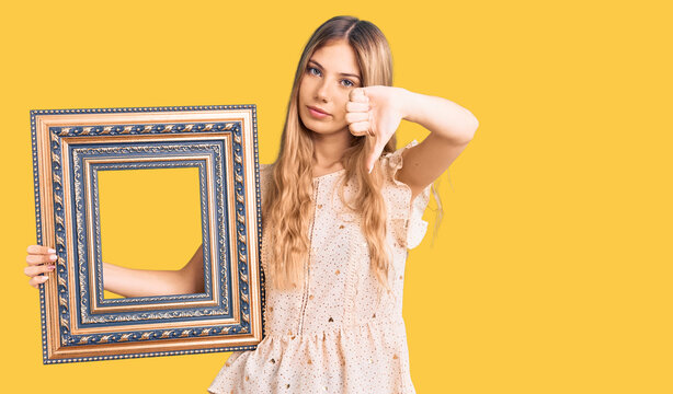 Beautiful caucasian woman with blonde hair holding empty frame with angry face, negative sign showing dislike with thumbs down, rejection concept