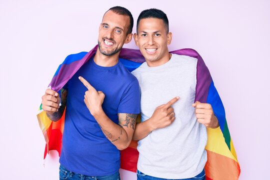 Young gay couple of two men holding rainbow lgbtq flag together smiling happy pointing with hand and finger