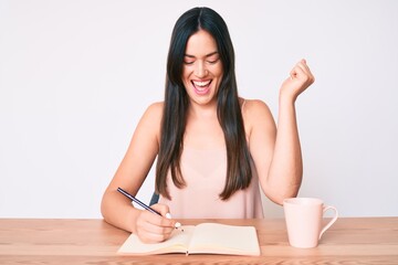 Young caucasian woman sitting at the desk writing book drinking coffee screaming proud, celebrating victory and success very excited with raised arms