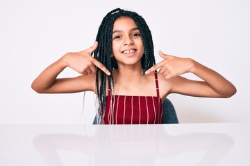 Young african american girl child with braids wearing casual clothes sitting on the table looking...