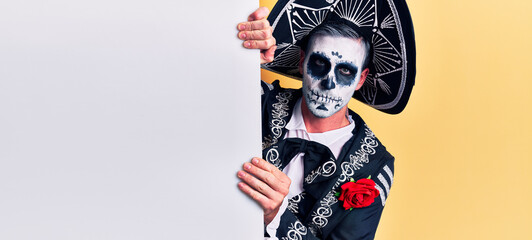 Young man wearing mexican day of the dead costume holding blank empty banner thinking attitude and sober expression looking self confident