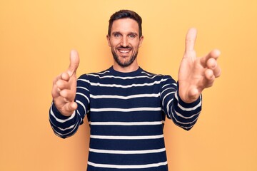 Young handsome man wearing casual striped sweater standing over isolated yellow background looking at the camera smiling with open arms for hug. Cheerful expression embracing happiness.