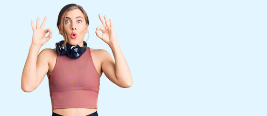 Beautiful caucasian young woman wearing gym clothes and using headphones looking surprised and shocked doing ok approval symbol with fingers. crazy expression