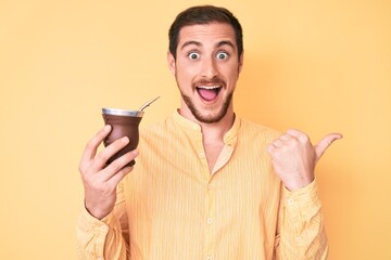 Young handsome man drinking mate infusion pointing thumb up to the side smiling happy with open...