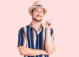 Young handsome caucasian man wearing summer hat with a big smile on face, pointing with hand and finger to the side looking at the camera.