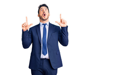 Young hispanic man wearing business clothes amazed and surprised looking up and pointing with fingers and raised arms.