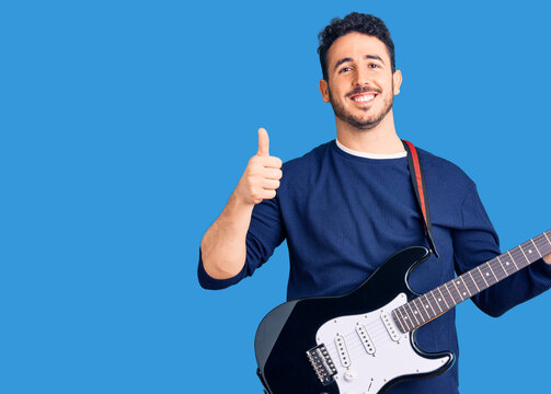 Young hispanic man playing electric guitar smiling happy and positive, thumb up doing excellent and approval sign