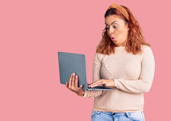 Young latin woman holding laptop scared and amazed with open mouth for surprise, disbelief face